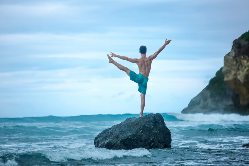 man practices yoga on a rock overlooking the ocean