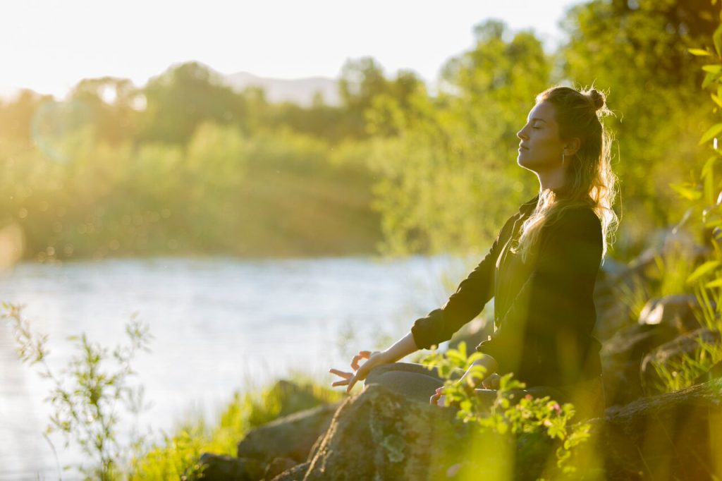 woman meditates near a river - yogatoday
